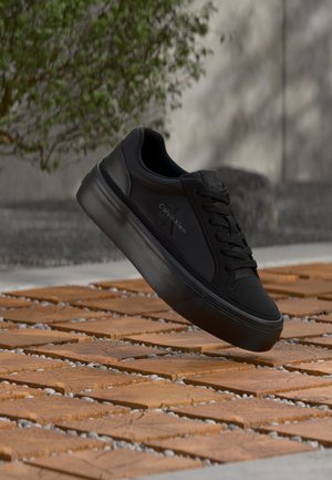 Baskets noires Calvin Klein à semelle épaisse suspendues au-dessus de dalles en bois en extérieur, avec un arrière-plan flou de verdure et de mur.