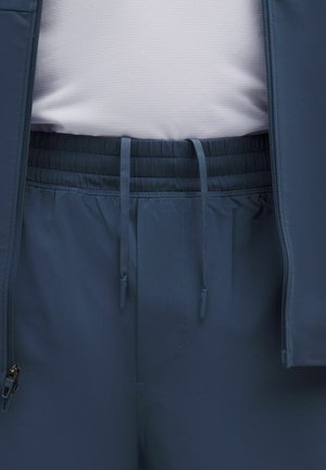 Veste de sport bleu marine avec une texture lisse et une taille élastique. Comprend une poche zippée et des cordons de serrage ajustables.