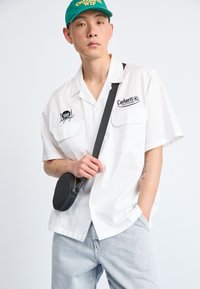 Camisa de botones de manga corta blanca con dos bolsillos frontales y bordado negro, que se lleva con unos jeans gris claro y un bolso cruzado negro redondo.