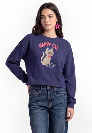 Kvinde iført en marineblå sweatshirt med en kattegrafik, teksten ""Happy Cat"" og blå jeans, poserer med den ene hånd på hoften og lyserøde stjerneøreringe.