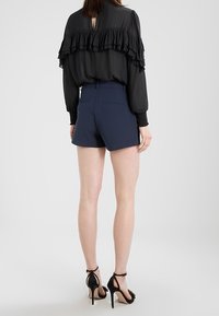 Žena v černé blůze s volány, modrých šortkách s vysokým pasem a černých sandálech na vysokém podpatku s páskem kolem kotníku, stojící zády k divákovi.