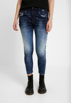 Personne portant un jean skinny bleu délavé avec des poches usées et des bottes de combat noires à lacets, debout sur un fond blanc.