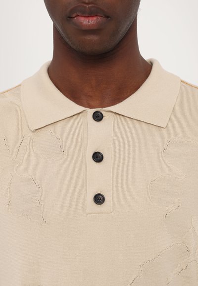 Beige stickad polo-tröja med ett texturerat blommönster, utrustad med en klassisk krage och tre svarta knappar längs placket.