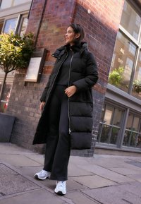 Giacca a vento nera con cappuccio, dotata di chiusura con zip e tasche laterali; indossata sopra un top nero e pantaloni svasati, abbinata a sneakers bianche.