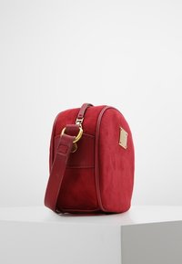 Rød ruskinds crossbody taske med afrundet form, detaljer i guldmetal og en enkelt justerbar rem. Glat tekstur med et logo-detalje på siden.