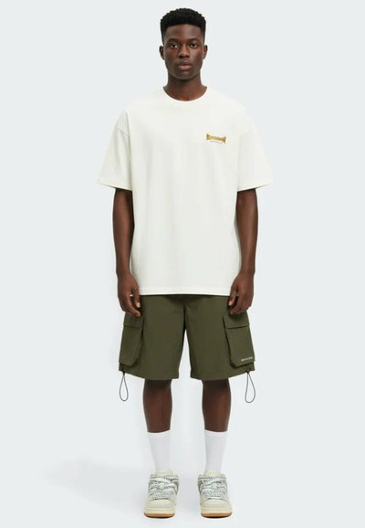 Giovane uomo in piedi, rivolto verso l'obiettivo, indossa una t-shirt bianca oversize, pantaloncini cargo verde oliva, calzini bianchi e sneakers su uno sfondo neutro.