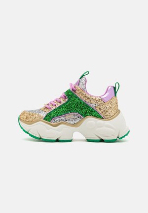 Zapatilla gruesa con paneles brillantes en dorado, verde y plata, cordones rosas, detalles satinados en púrpura y una suela blanca gruesa con dibujo verde.