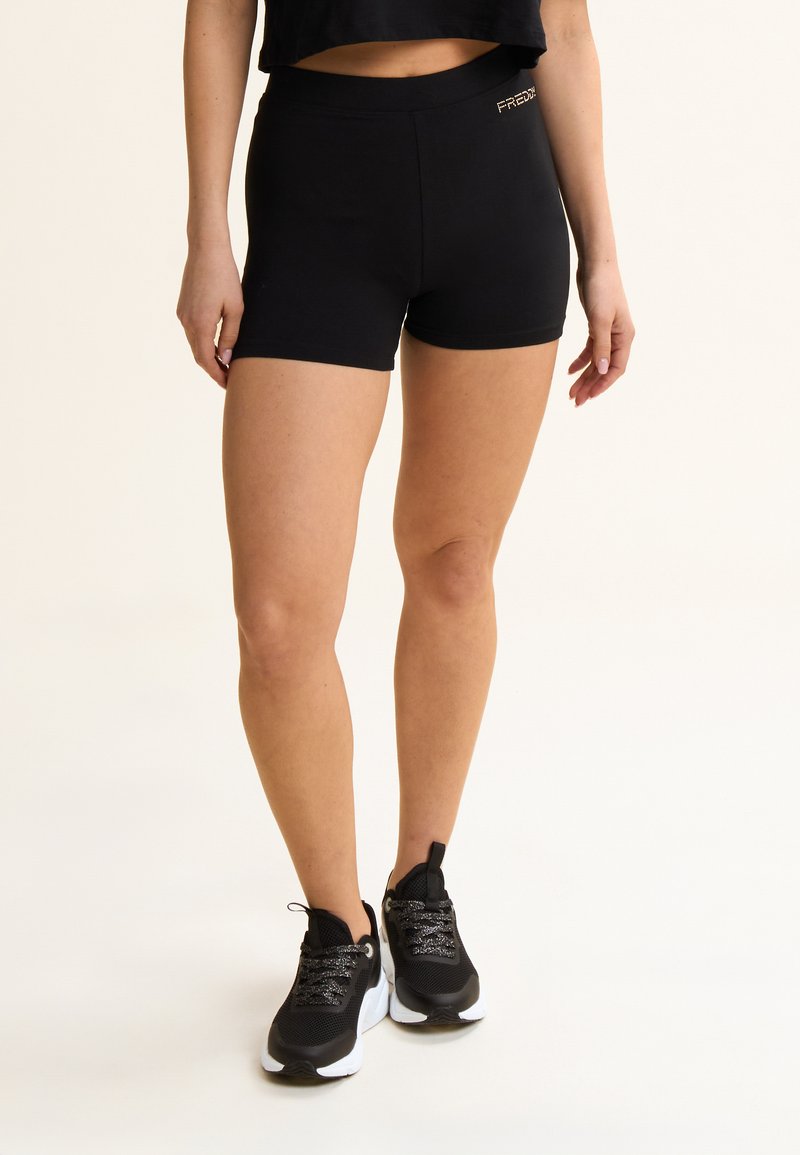 Shorts de sport noirs en tissu lisse avec une coupe ajustée. Dotés d'une taille élastique et d'un petit logo doré en accent. Portés avec des baskets noires.
