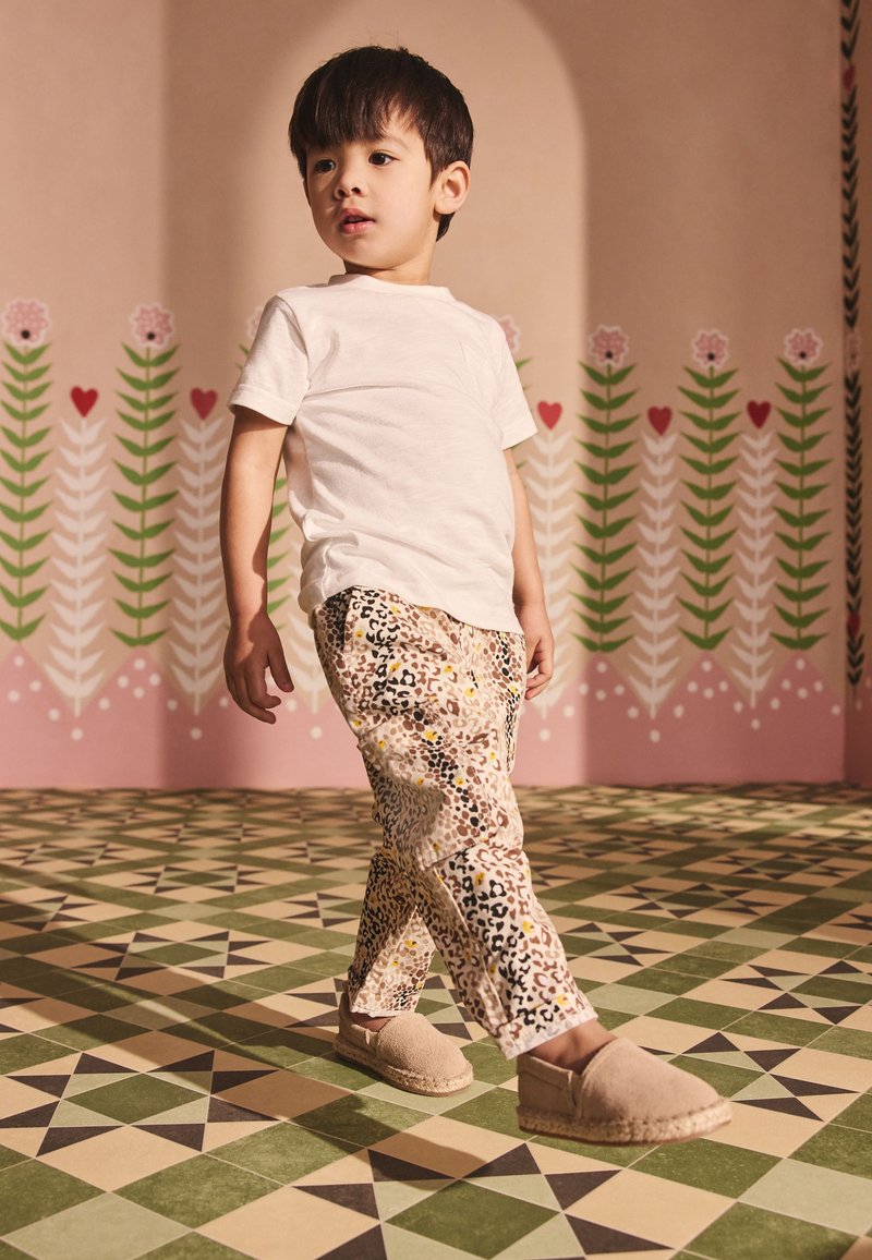 Jeune garçon marchant à l'intérieur portant un t-shirt blanc, un pantalon à motifs et des chaussures beiges sans lacets sur un fond floral et géométrique.