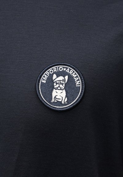 Στρογγυλό κεντητό μοτίβο σε μαύρο ύφασμα με εικονογράφηση καθισμένου σκύλου και το κείμενο "EMPORIO ARMANI" γύρω από την άκρη.