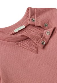 Zoom su un colletto di una maglietta lavorata a maglia di un rosa polveroso, con bottoni a pressione metallici e un'etichetta di abbigliamento bianca visibile.