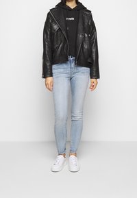 Chaqueta de cuero negra estilo motero con acabado texturizado, llevada sobre una sudadera con capucha negra. Jeans ajustados azul claro y zapatillas blancas completan el conjunto.