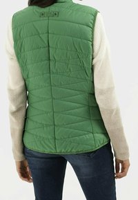 Gilet matelassé vert avec un col montant, présentant un motif en chevrons. Manches longues crème en tricot texturé. Porté sur un jean foncé.