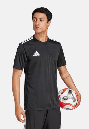 Mann in schwarzer Adidas-Sportbekleidung hält einen rot-weißen Fußball und schaut leicht nach links vor weißem Hintergrund.