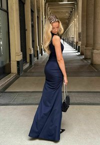 Tmavě modré, přiléhavé, bezramínkové večerní šaty s dlouhou sukní a řaseným designem. Modelka drží černou kabelku s řetízkovým popruhem.