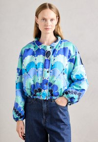 Giacca stampata blu e verde con motivo a conchiglia, vita elastica e bottoni a pressione, abbinata a jeans in denim scuri.