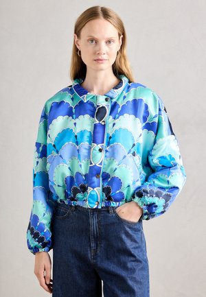 Giacca stampata blu e verde con motivo a conchiglia, vita elastica e bottoni a pressione, abbinata a jeans in denim scuri.