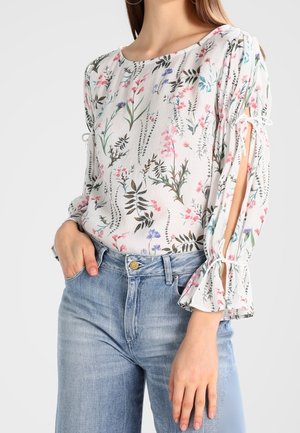 White long-sleeve blouse with pink, purple, and green floral print, featuring tied slits on sleeves, paired with light blue high-waisted jeans.