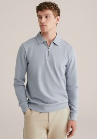 Light blue long-sleeve polo shirt with a zippered collar, ribbed cuffs, and a soft texture. Worn with beige trousers for a casual look.