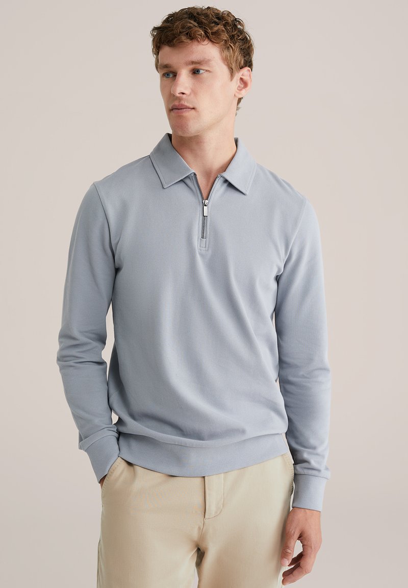 Light blue long-sleeve polo shirt with a zippered collar, ribbed cuffs, and a soft texture. Worn with beige trousers for a casual look.