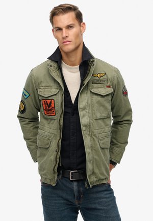 Chaqueta verde oliva con múltiples parches y bolsillos, con cremallera, cuello y un corte relajado, llevada sobre una camiseta negra.