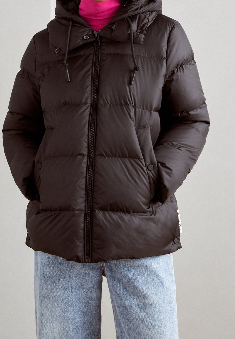 Veste matelassée noire avec capuche, fermeture éclair à l'avant et cordons de serrage. Comprend un rembourrage cousu, des poches latérales et une coupe décontractée. Portée avec un jean bleu.