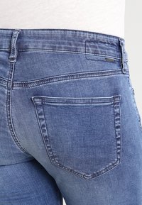 Mörkblå denimjeans med fem fickor, synlig sömnad och en märkesetikett på baksidan av midjebandet. Tyget har en ljus tvättstruktur.