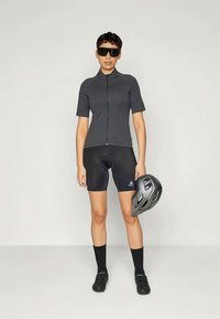 Koszulka kolarska z krótkim rękawem, zapinana na zamek, w odcieniu szarym, noszona z czarnymi szortami. Czarne kask w jednej ręce, widać buty kolarskie oraz skarpetki do kostek.