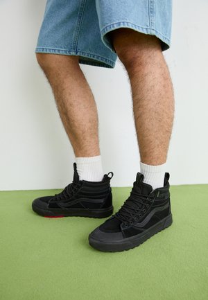 Ben i denimshorts iført hvide mellemlange strømper og sorte høje sneakers på grøn gulv mod hvid væg.