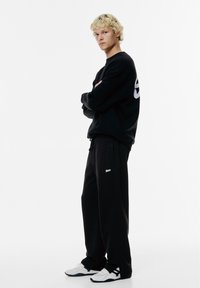 Zwarte sweatshirt met groot logo op de achterkant, gecombineerd met zwarte sweatpants met een logo. Witte sportschoenen met zwarte accenten.