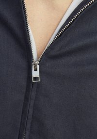 Tumesinane tõmbetaskuga jope, millel on metallne tõmblukk, tekstuurne kangas ja valge vooder, mis on nähtav krae juures.