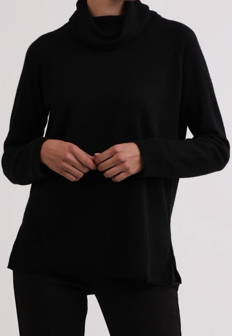 Personne portant un pull à col roulé noir à manches longues, debout les mains doucement jointes devant un fond uni.