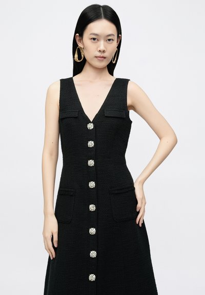 Melnas tweeda kleita ar V veida kakla izgriezumu, pieciem dekoratīviem pogām, divām priekšējām kabatām un volānu akcentiem. Modelis valkā lielas zelta riņķa auskarus.