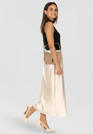 Vrouw glimlachend, gekleed in een zwart mouwloos topje, een crèmekleurige satijnen midirok, beige hakken, en draagt een kleine beige handtas over haar schouder.