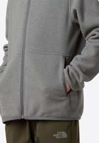 Sivá fleecová bunda s vreckami na zips a hladkou textúrou. Má vysoký golier a elastické manžety. Kombinovaná s tmavozelenými nohavicami.