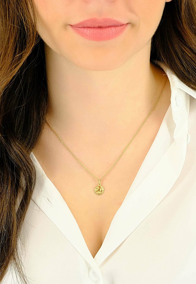 Collier pendentif en or sur une chaîne délicate, avec un charme circulaire orné de motifs complexes, porté sur une chemise blanche à col.