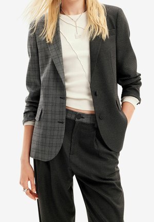 Kobieta ubrana w dwukolorową szarą kratkę marynarkę i pasujące spodnie, nałożone na biały krótki prążkowany top z srebrnym naszyjnikiem w kształcie serca.