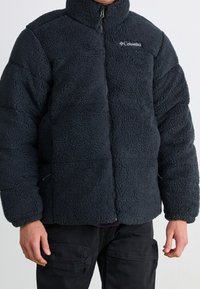 Tmavě šedá fleecová bunda s vysokým límcem, zapínáním na zip a texturovaným povrchem. Na hrudi je logo a bunda má pevnou, polstrovanou konstrukci.