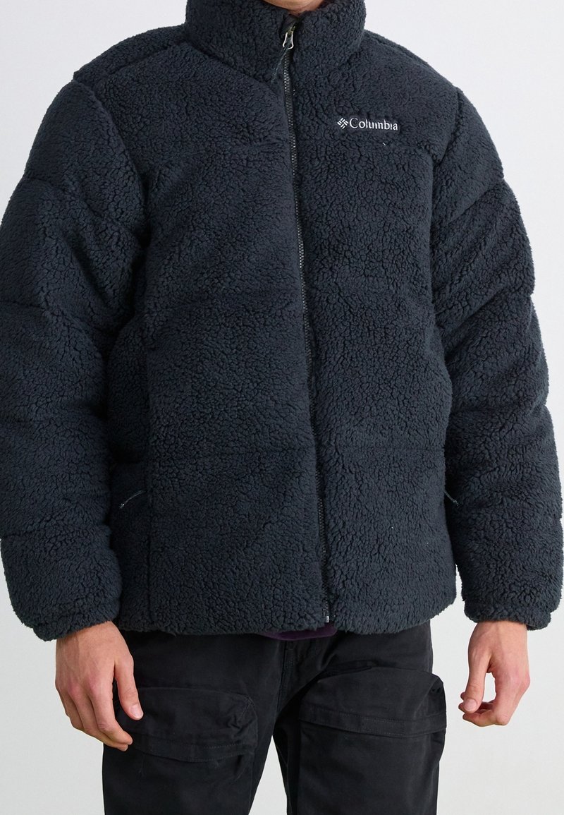 Tmavě šedá fleecová bunda s vysokým límcem, zapínáním na zip a texturovaným povrchem. Na hrudi je logo a bunda má pevnou, polstrovanou konstrukci.