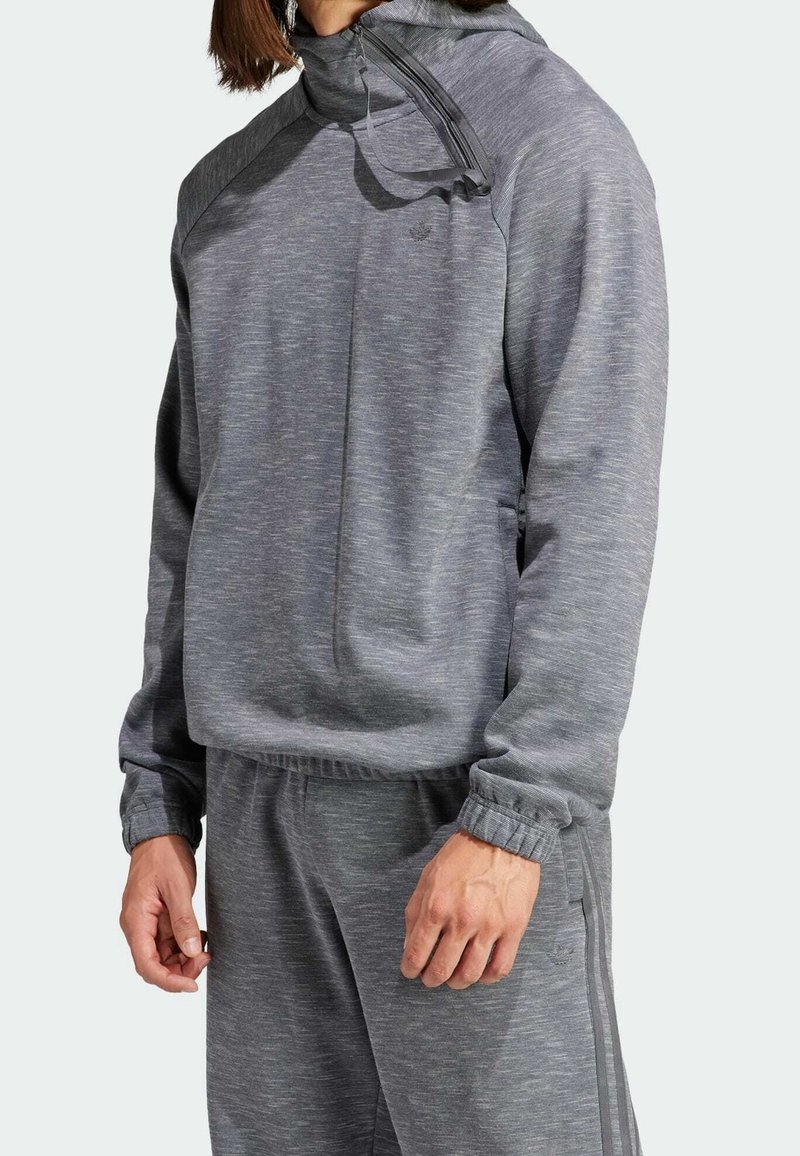 Man som bär en ljusgrå träningsjacka med hög krage och dragkedja samt matchande träningsbyxor, stående mot en neutral bakgrund.