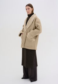 Cappotto in pelliccia sintetica beige con colletto a scialle, ampie tasche e vestibilità morbida, indossato sopra un lungo vestito di maglina marrone e pantaloni scuri.