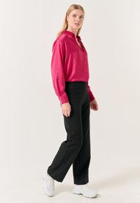 Fuchsia knopjesblouse met een ontspannen pasvorm, gecombineerd met zwarte rechte broek en witte sneakers, tegen een neutrale achtergrond.