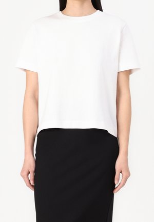 Femme portant un t-shirt blanc uni à manches courtes et une jupe noire ajustée, face à un fond blanc.