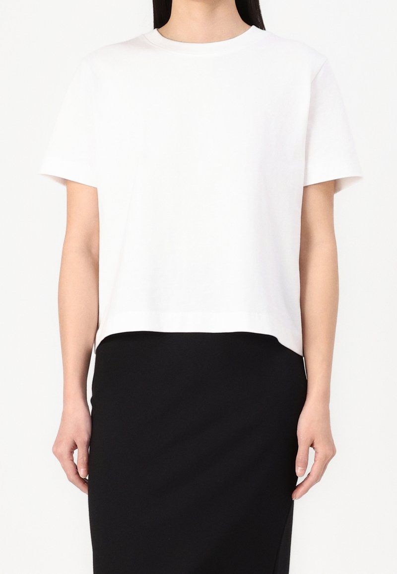 Femme portant un t-shirt blanc uni à manches courtes et une jupe noire ajustée, face à un fond blanc.