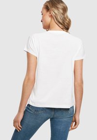 Bílý krátký rukáv T-shirt, měkký materiál, uvolněný střih, má kulatý výstřih a jemné prošívání. V kombinaci s modrými džíny.