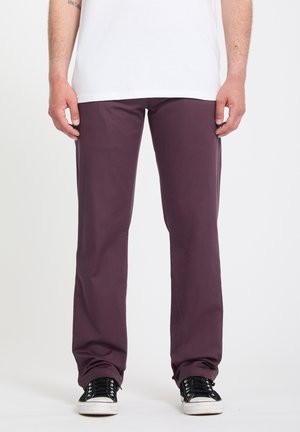 Άνδρας που φοράει παντελόνι σε χρώμα δαμάσκηνου με ίσια γραμμή, μαύρα αθλητικά με λευκές σόλες, και λευκό μπλουζάκι, στέκεται μπροστά σε απλό φόντο.