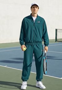 Mladý muž v tyrkysovej teplákovej súprave a bielej šiltovke drží tenisovú loptičku a raketu na vonkajšom tenisovom kurte.