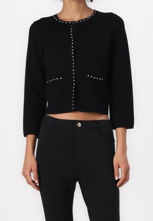 Nainen, joka käyttää istuvaa mustaa lyhytmallista neuletakkia, jossa on hopeisia niittejä kaula-aukon, napituslinjan ja taskujen ympärillä, yhdistettynä mustiin korkeavyötäröisiin housuihin.