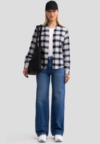 Schwarz-weiß kariertes Flanellhemd, weißes T-Shirt, blaue weite Jeans, schwarze Mütze und schwarze Tragetasche; weiße Sneakers.