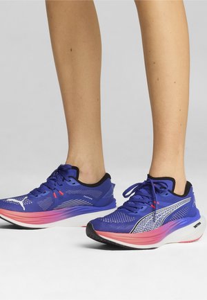 Person wearing blue and pink athletic running shoes standing on a light gray surface with legs visible from mid-calf down.